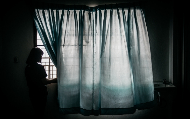 Young woman backlit, pulling back curtain and looking out window